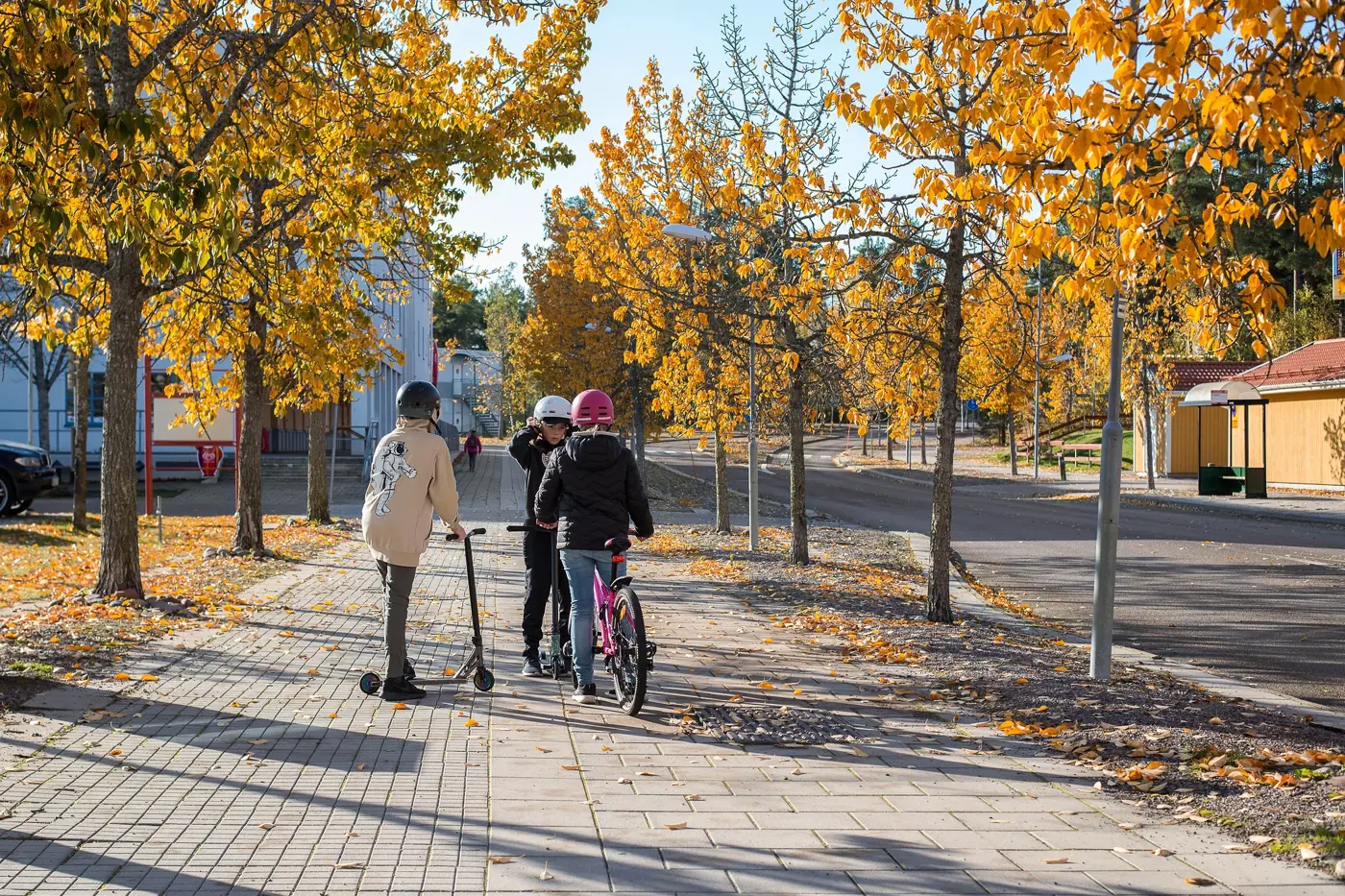 Tre barn med hjälm på huvudet står och pratar på en gång-och cykelbana. Två står med varsin kickbike och den tredje med en cykel.