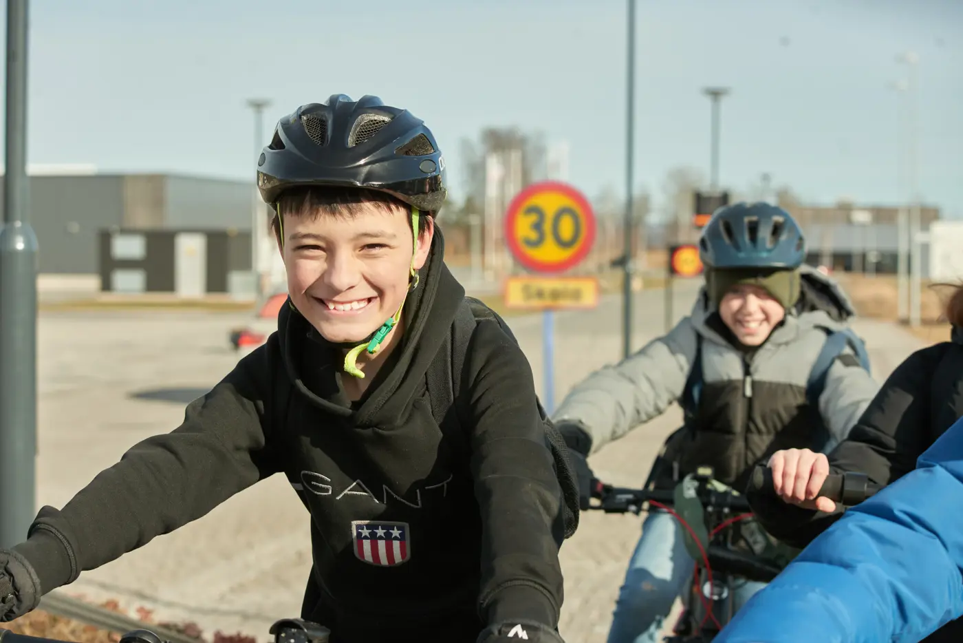 En grupp skolbarn kommer cyklandes och ler mot kameran. Två syns med ansiktet i bild och båda har cykelhjälm på huvudet.